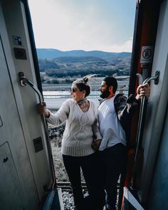 a man and woman standing on the side of a train car with mountains in the background