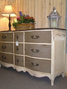 an old dresser with flowers on top and a lamp next to it in front of a wood paneled wall