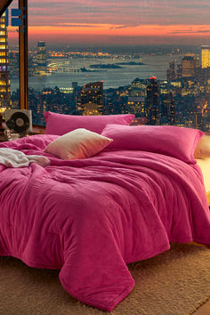 a bed with pink comforter and pillows in front of a window overlooking the city