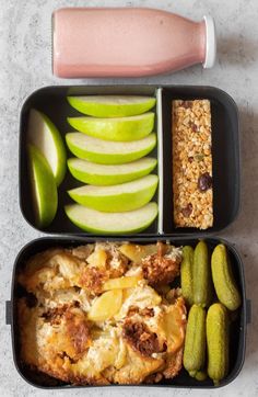 two black trays filled with different types of food next to pickles and apple slices