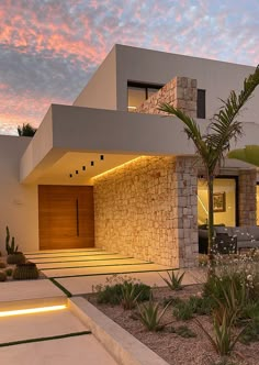 a modern house is lit up at night with lights on the side and palm trees in front