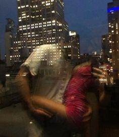 two people hugging each other in front of the city skyline at night with skyscrapers lit up behind them