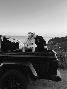 two people sitting in the back of a truck near the ocean and mountains, one person is holding his arm around the other man's shoulder