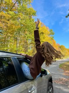 a woman is waving out the window of a car on a road with trees in the background