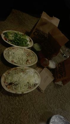 three plates of food sitting on the floor next to a bag of chips and salad