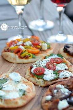 several small pizzas with different toppings are on a wooden board next to wine glasses