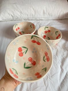 three bowls with cherries painted on them are sitting on a bed and someone is holding the bowl in their hand