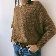 a woman standing in front of a white wall with her hands on her hips wearing a brown sweater and jeans