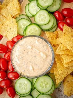 a bowl of dip surrounded by cucumbers, tomatoes and chips on a cutting board