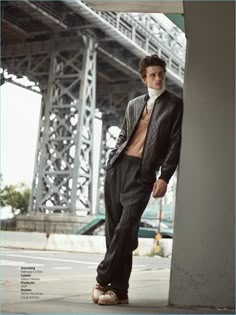 a young man leaning against a wall in front of a large metal structure wearing a suit and tie