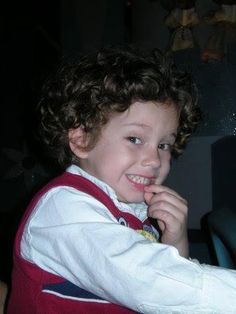 a young boy sitting in front of a laptop computer smiling at the camera while wearing a red vest