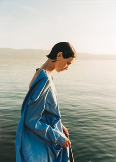 a woman standing on the edge of a body of water wearing a blue dress and carrying a brown bag