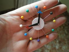 a hand holding a small clock with pins on it's face and multicolored nails