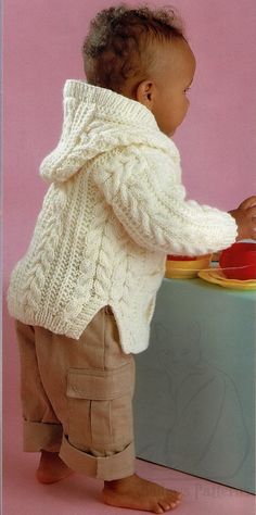 a baby in a sweater and pants standing at a table with a plate on it