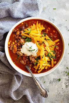 a white bowl filled with chili and cheese on top of a blue towel next to a spoon