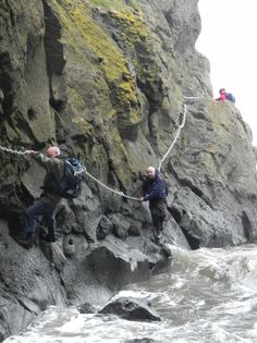 three people climbing up the side of a cliff by the ocean with ropes attached to them