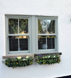 two windows with shutters on the side of a white building, one has flowers in it