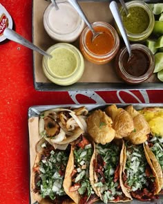 two trays filled with tacos and condiments next to each other on a red table