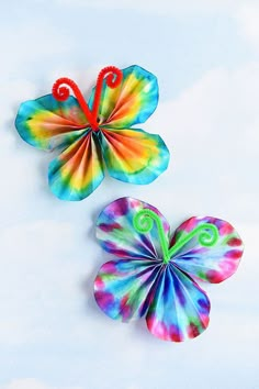 two colorful paper butterflies sitting next to each other on a white surface with clouds in the background