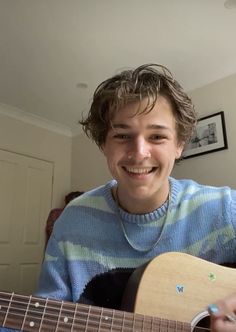 a young man smiles as he plays an acoustic guitar in his living room with a blue and white striped sweater