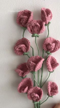 crocheted pink flowers with green stems are displayed on a white wall in front of a person's hand