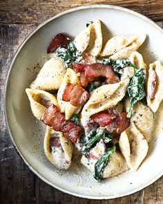 a white bowl filled with pasta and bacon on top of a wooden table next to a fork