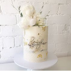 a wedding cake decorated with white flowers and gold lettering on a stand against a brick wall