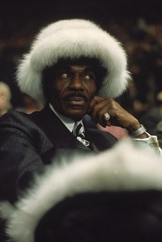 a man wearing a white fur hat sitting in front of an audience with his hand on his chin