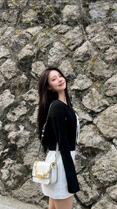 a woman standing in front of a rock wall holding a white purse and smiling at the camera