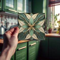 a hand holding up a green and white tile in front of a kitchen counter top