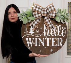 a woman holding up a wooden sign that says hello winter with a bow on it