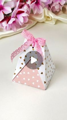 a small pink and white box with a bow on the top sitting next to purple flowers