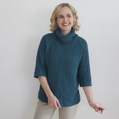 a woman standing in front of a white wall wearing a green sweater and beige pants