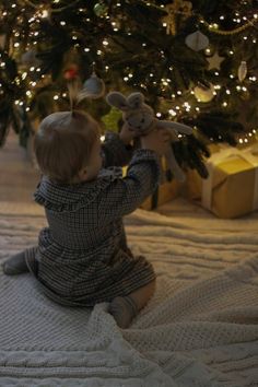 a baby sitting on the floor playing with a teddy bear next to a christmas tree