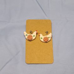 two small white and green chicken earrings on a brown piece of paper next to a blue background