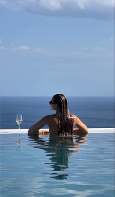 a woman sitting in the water with a glass of wine next to her and looking out at the ocean