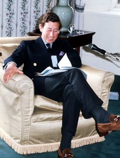 a man in a suit sitting on a chair reading a book and smiling at the camera