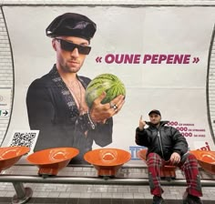 a man is sitting on a bench holding a watermelon in front of a poster