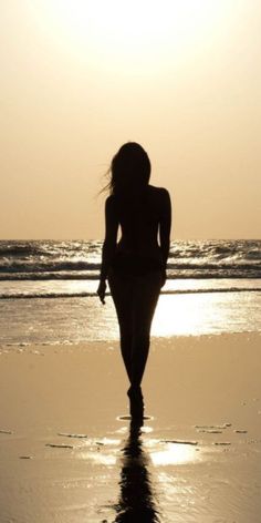 a woman walking on the beach at sunset with her back to the camera, silhouetted by the sun