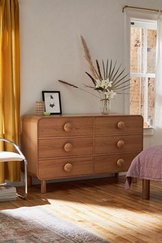 a bedroom scene with focus on the dresser and bed in the foreground, and sunlight coming through the window