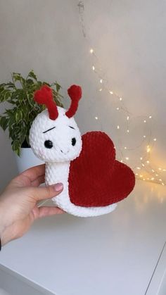 a hand holding a small stuffed animal with a heart on it's chest, next to a potted plant