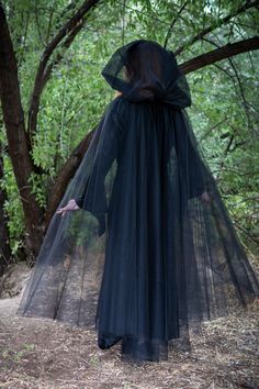 a woman in a black dress and veil walking through the woods with her hands on her hips