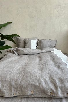 an unmade bed with grey linens and white pillows, next to a potted plant