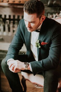 a man in a tuxedo sitting on a chair looking down at his watch