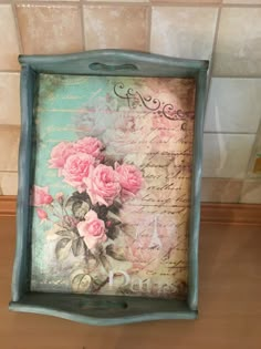 a blue frame with pink roses on it sitting on top of a wooden table next to a tile wall