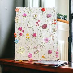 an open book with flowers on it sitting on a table next to a window and a cell phone