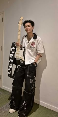 a man in black pants and white shirt holding an electric guitar while standing next to a wall