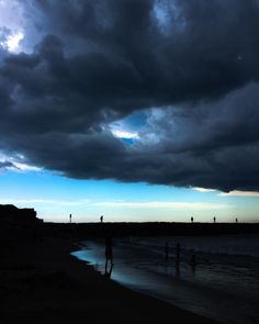 some people are standing on the beach under a dark sky with clouds in the background