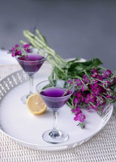 two glasses filled with purple liquid sitting on top of a white plate next to flowers