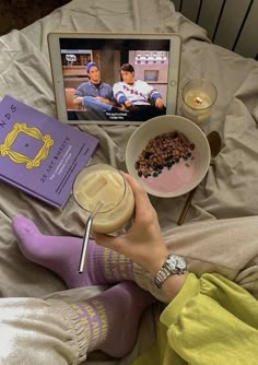 a person laying on a bed with food and a book in front of them while watching tv